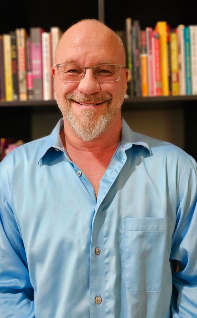 Picture of D. Woodja Flanigan from waist up, in blue shirt, in front of bookcase.